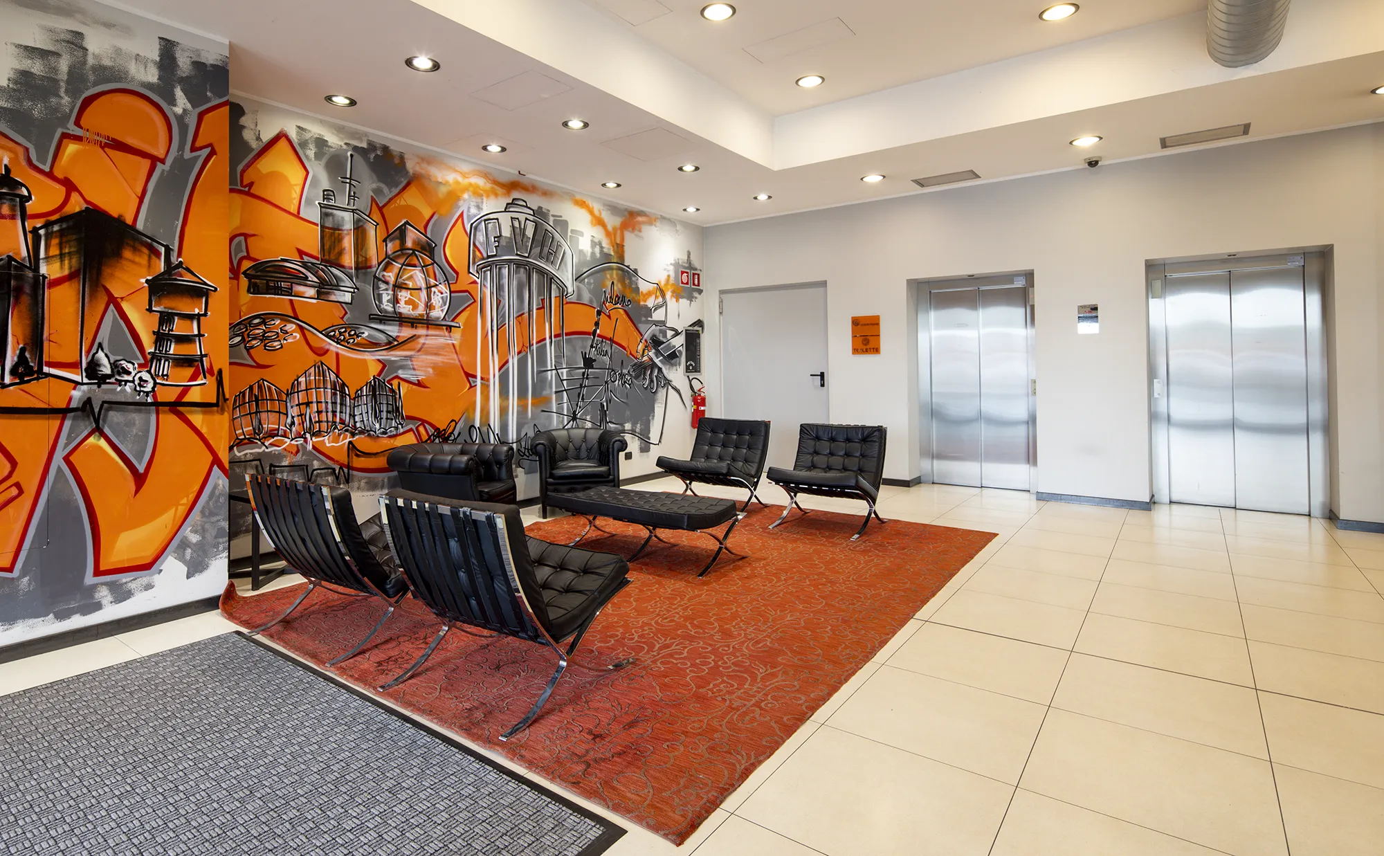 Nell'immagine, la Reception del Falck Village Hotel: si vede un tavolino con alcune poltroncine nere. La parete a sinistra è decorata da un murales sui toni dell'arancione. Di fronte, due ascensori.