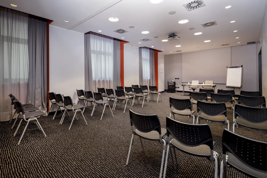 Nell'immagine, la sala meeting Unione del Falck Village Hotel da una prospettiva laterale: si vedono alcune file di sedie e, di fronte, la scrivania dei relatori e un lavagna a fogli mobili. Sulla sinistra, alcune finestre.